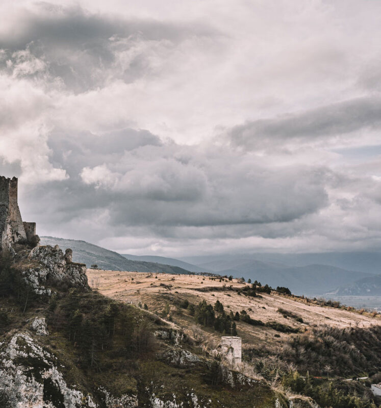 Karge hügelige Landschaft und Burgruine am linken Rand des Bildes mit kontrastreichen, grauen Wolken im Hintergrund.