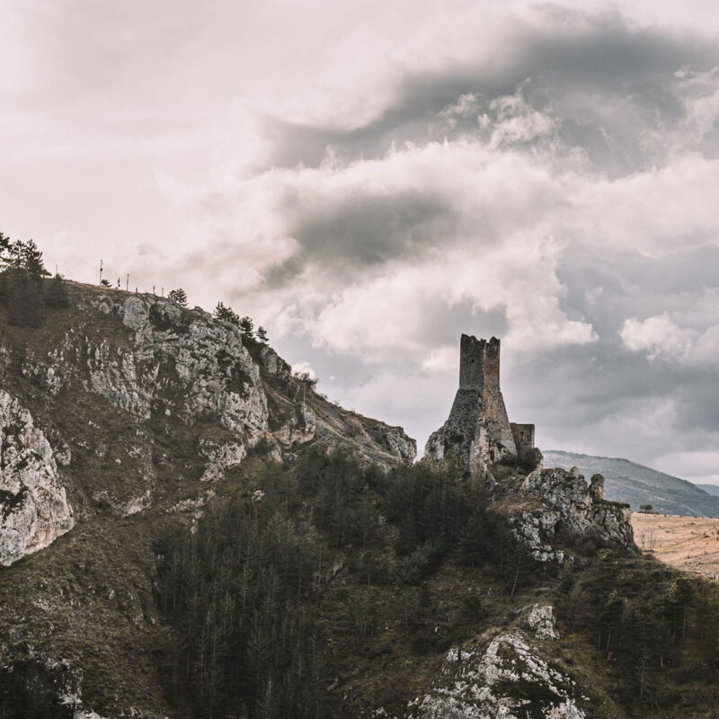 Karge hügelige Landschaft und Burgruine in der rechten Hälfte des Bildes mit kontrastreichen, grauen Wolken im Hintergrund.